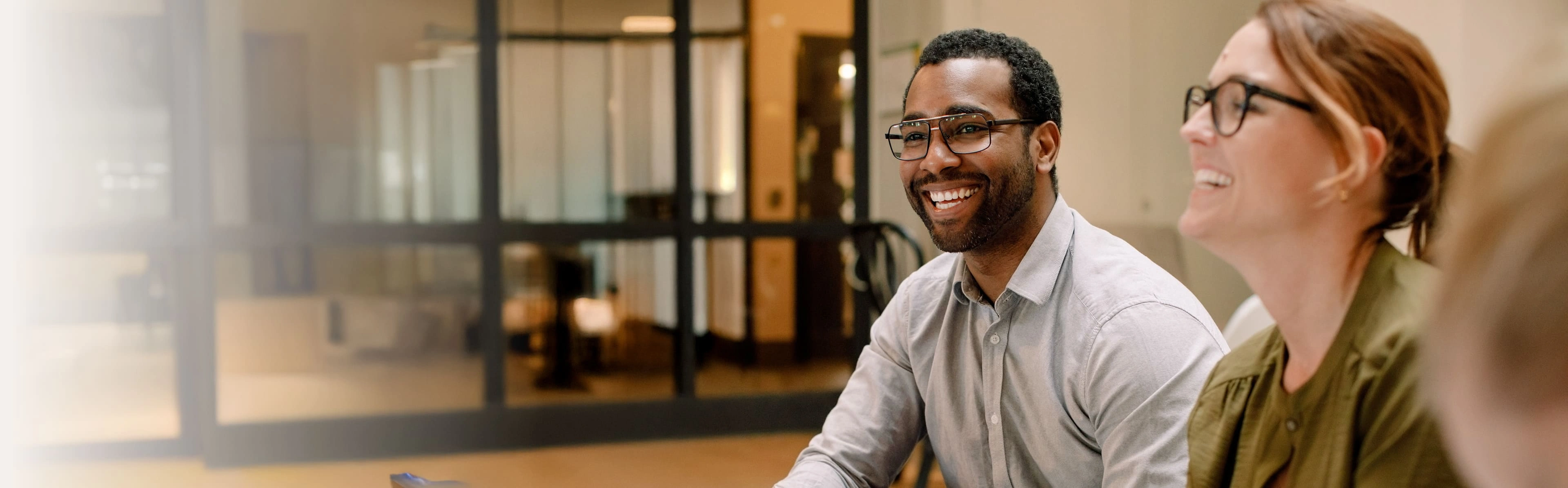 Indoor meeting with three people talking in a modern room with warm lighting. One person holds a white mug, another wears glasses and a light shirt, and the third has tied-back hair. Glass doors and cozy interior design in the background.
